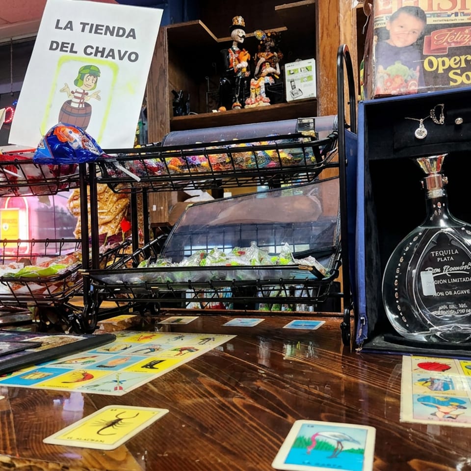 Mexican market counter displaying colorful candies, lottery cards, magazines, figurines, and a boxed tequila bottle.